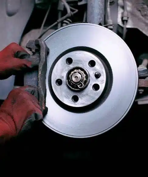A person wearing red gloves is installing or inspecting a car’s brake disc and caliper. The image shows a close-up of the vehicle’s exposed wheel hub and brake assembly.